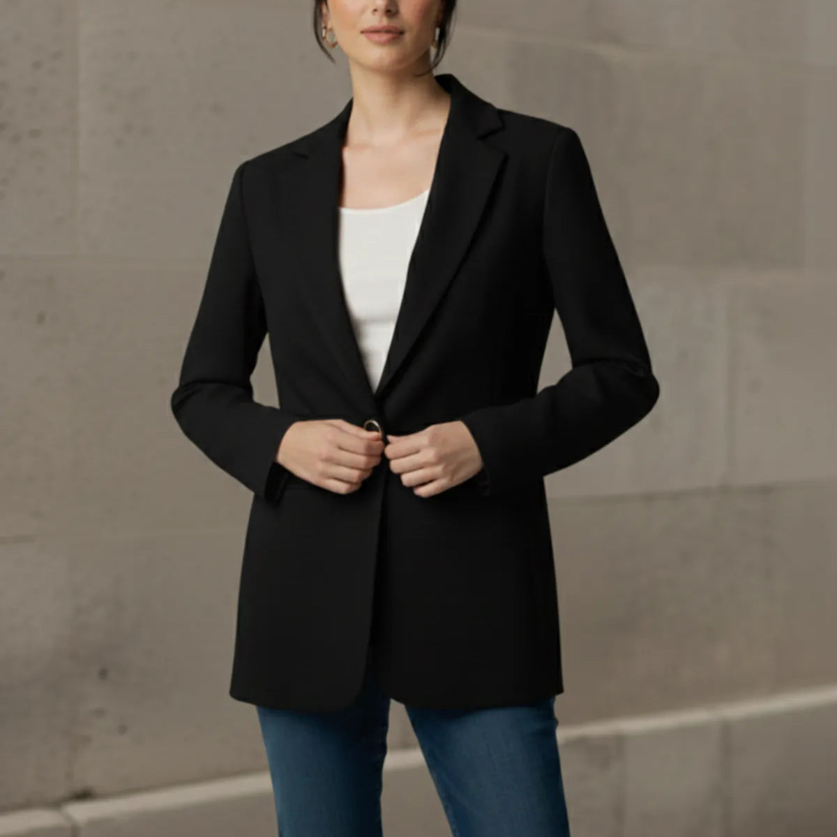 Woman wearing a black blazer over a white top and blue jeans against a beige wall.
