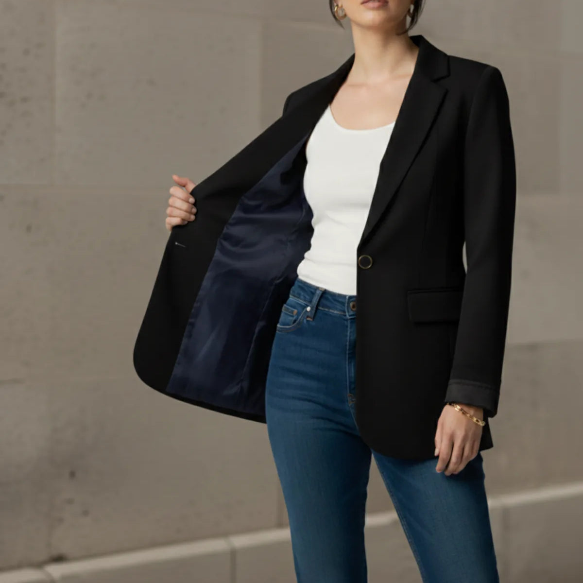 Woman wearing a black blazer, white top, and blue jeans against a concrete wall.