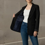 Woman wearing a black blazer, white top, and blue jeans against a concrete wall.