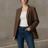Woman wearing a mocha blazer, white top, and blue jeans against a stone wall.