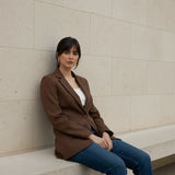 Woman sitting on a bench against a beige wall wearing a mocha blazer 