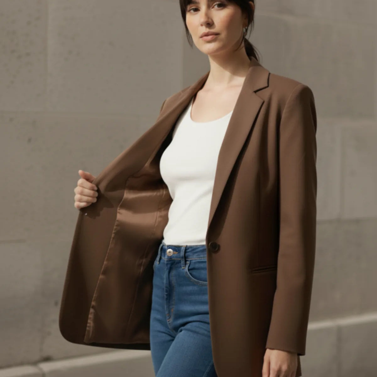 Woman wearing a mocha blazer over a white top and blue jeans against a stone wall.