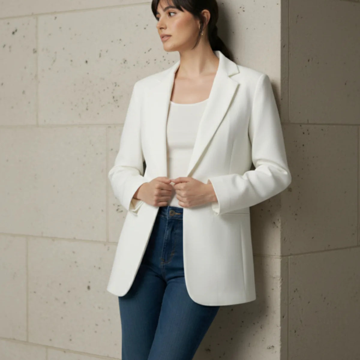 Woman wearing a white blazer and blue pants against a light-colored wall.