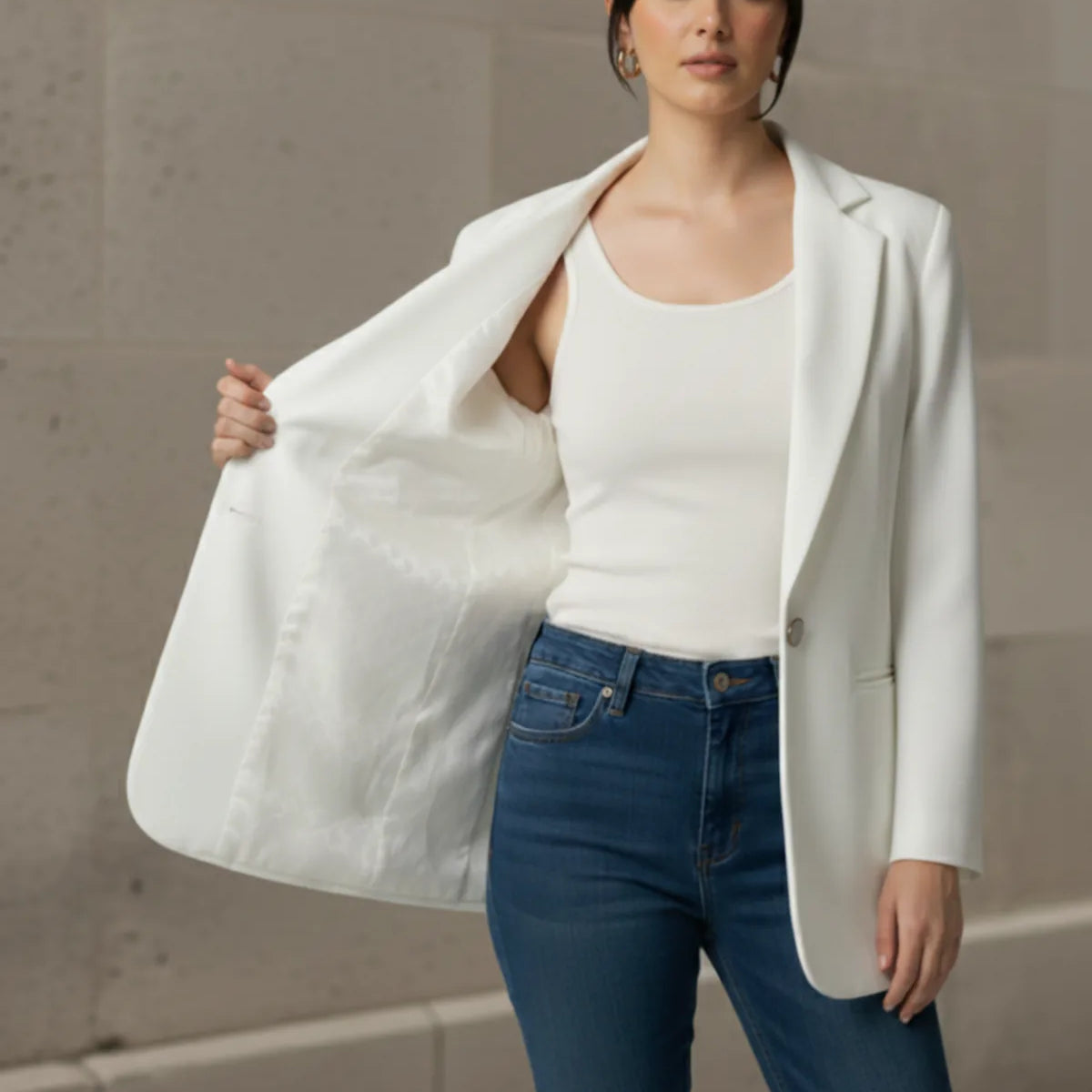 Woman wearing a white blazer over a white top and blue jeans against a neutral wall.