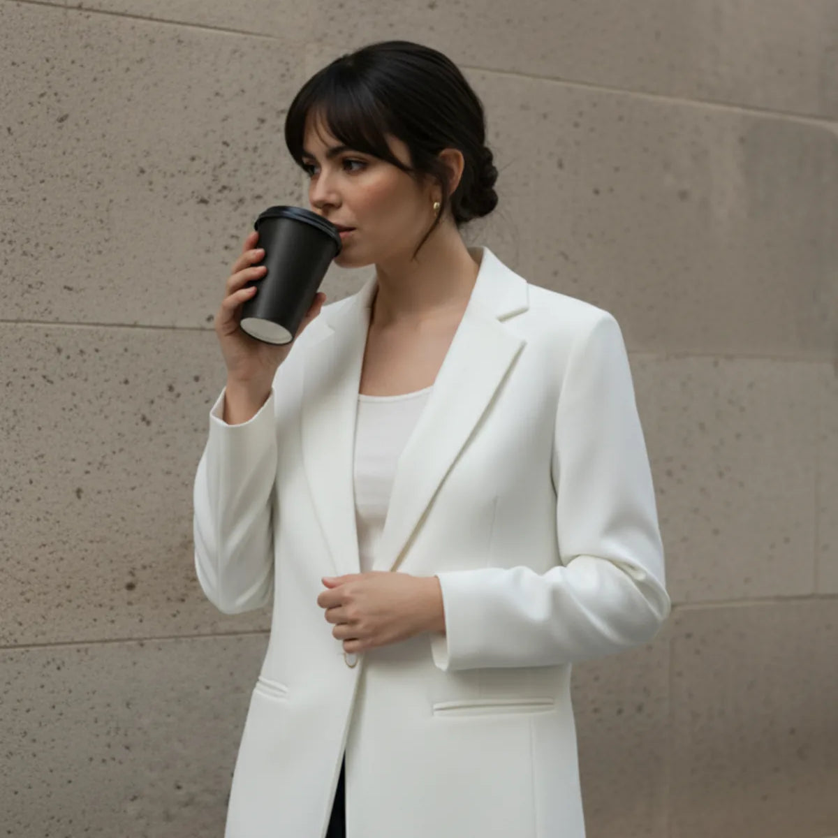 Woman in a white blazer drinking from a black cup against a beige wall.