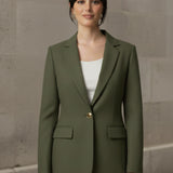 Woman wearing an Olive blazer over a white top and blue jeans against a stone wall.