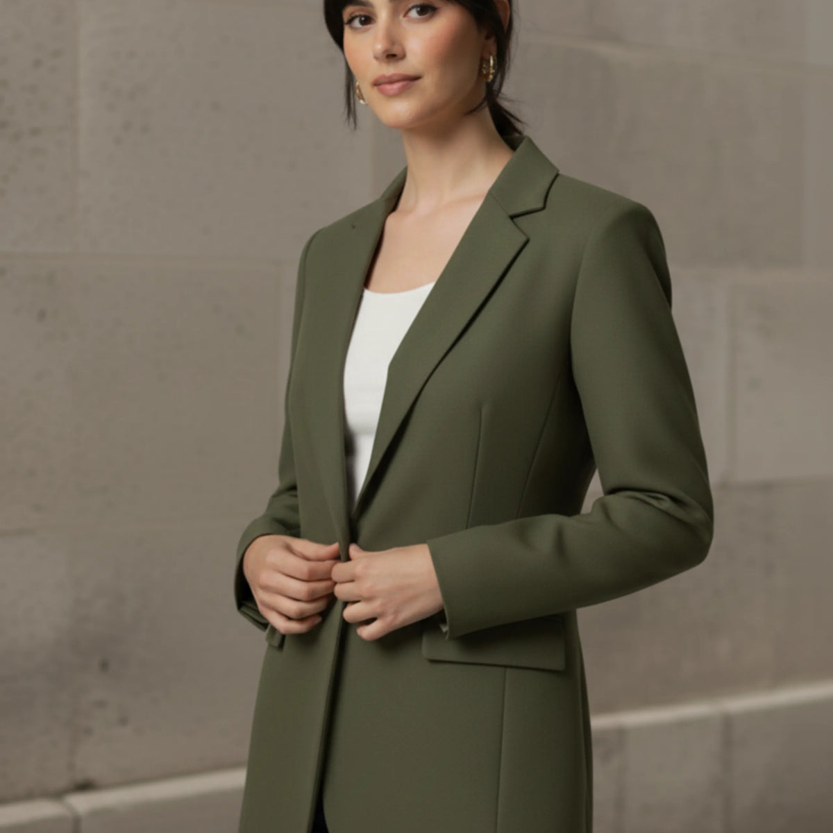Woman wearing an olive blazer over a white shirt with blue jeans against a stone wall.