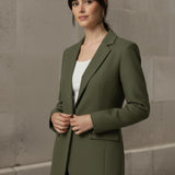 Woman wearing an olive blazer over a white shirt with blue jeans against a stone wall.