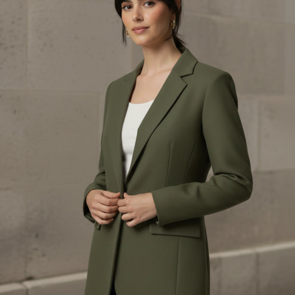 Woman wearing an olive blazer over a white shirt with blue jeans against a stone wall.