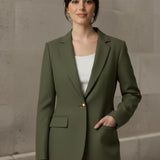 Woman wearing an olive blazer over a white top and blue jeans against a stone wall.