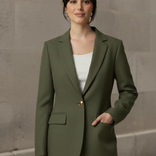 Woman wearing an olive blazer over a white top and blue jeans against a stone wall.