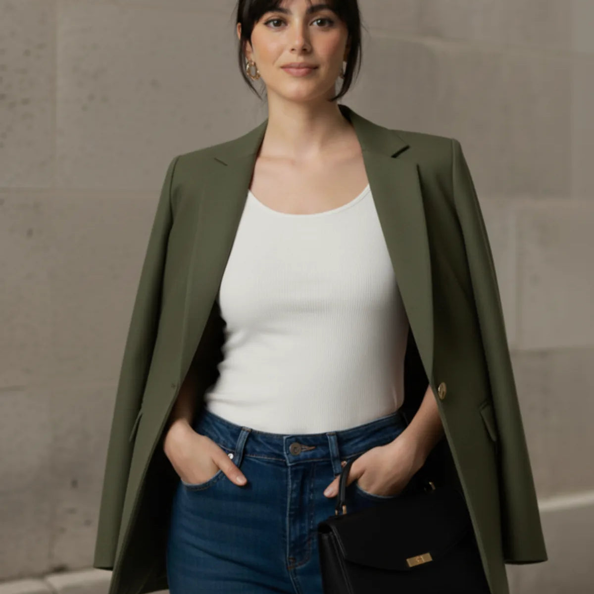 Woman wearing an olive blazer, white top, and blue jeans, standing against a stone wall.