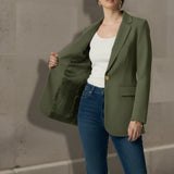 Woman wearing an olive blazer over a white top and blue jeans against a concrete wall.
