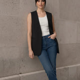 Woman leaning against a stone wall modeling a chic black longline sleeveless vest, styled casually with a white tank top and blue jeans.