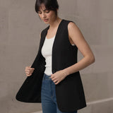 Woman adjusting the front hem of a black longline sleeveless vest, displaying the open-front drape and styling details.