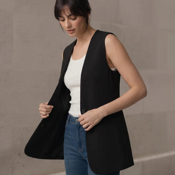 Woman adjusting the front hem of a black longline sleeveless vest, displaying the open-front drape and styling details.
