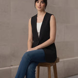 Woman seated on a wooden stool wearing a versatile black sleeveless longline blazer paired with denim jeans.
