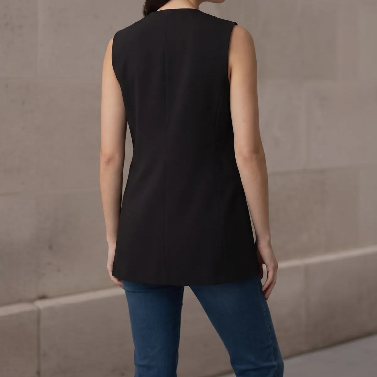 Back view of a woman wearing a tailored black sleeveless vest, showcasing the clean back seams, structure, and longline length.