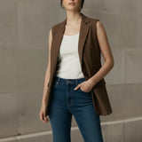 Woman standing casually with one hand in her jeans pocket, wearing a stylish open-front brown sleeveless blazer layered over a white tank top.