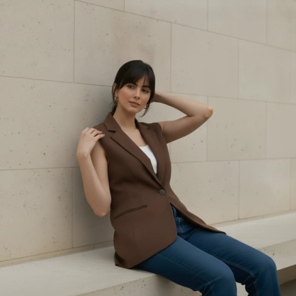 Woman sitting elegantly on a ledge with her hand near her hair, dressed in a sophisticated brown sleeveless longline blazer and casual jeans.