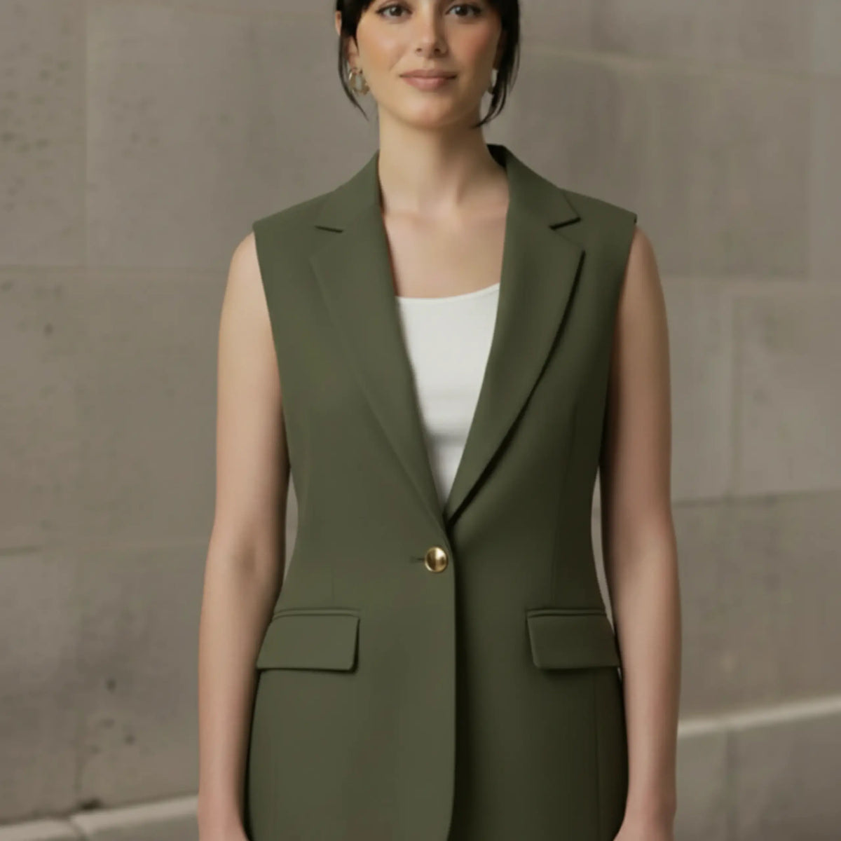 Front view of a woman modeling an olive green tailored sleeveless blazer fastened with a single gold button, featuring classic lapels and flap pockets.