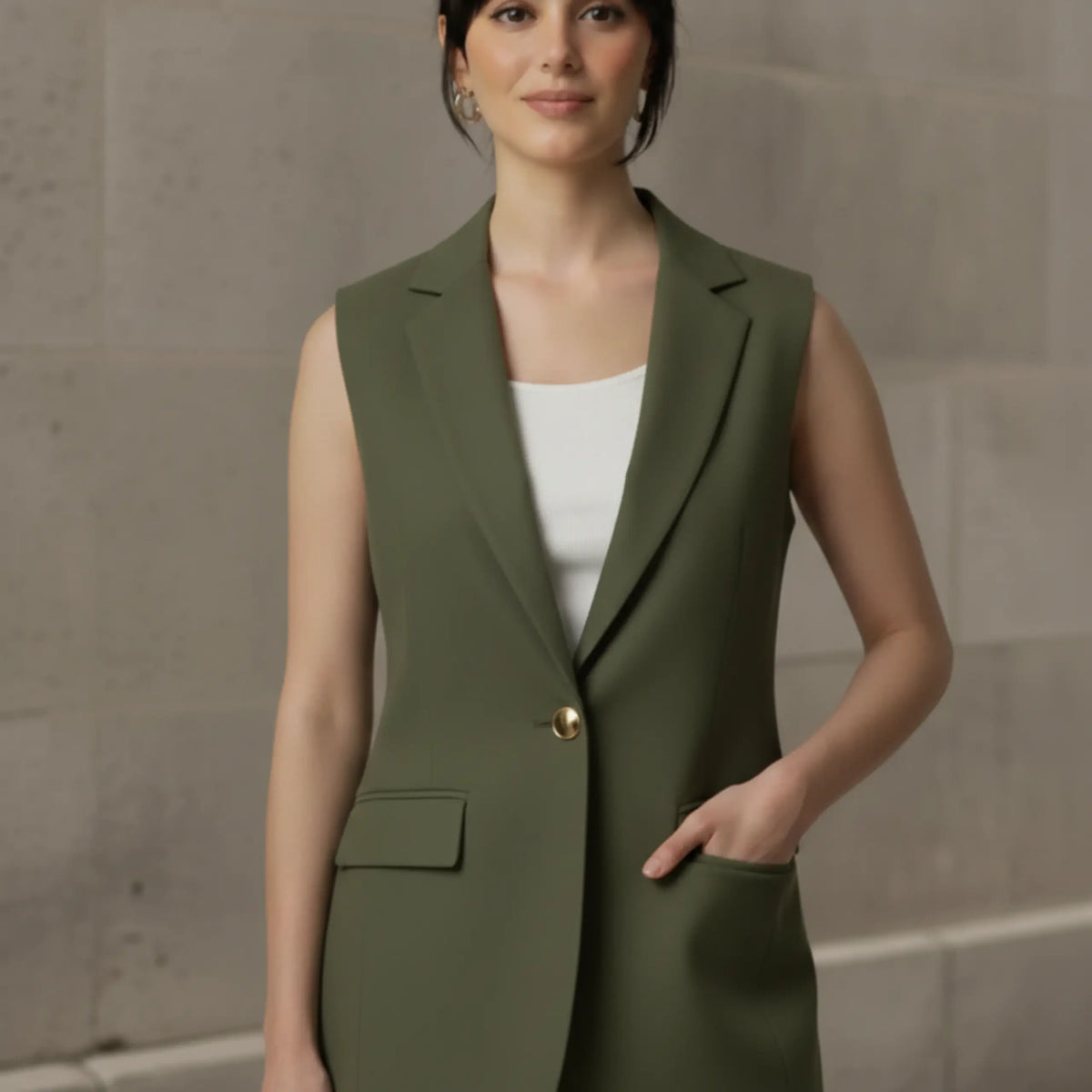 Woman standing with one hand in the front flap pocket of a buttoned olive green sleeveless blazer, showcasing the functional design and structured silhouette.