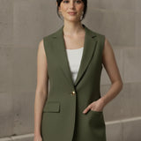 Woman standing with one hand in the front flap pocket of a buttoned olive green sleeveless blazer, showcasing the functional design and structured silhouette.