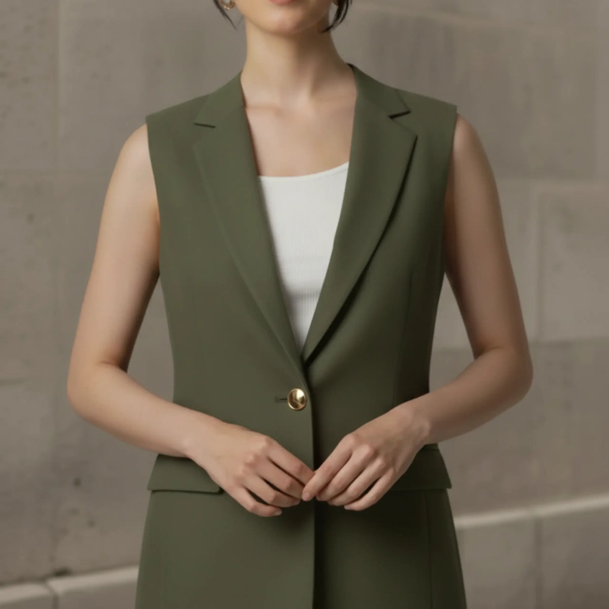 Close-up on the torso of an olive green tailored sleeveless vest worn over a white top, emphasizing the crisp lapels, smooth fabric texture, and statement gold button closure.