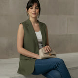 Lifestyle shot of a woman seated elegantly while wearing an open olive green sleeveless blazer, styled casually with blue denim jeans and a beige handbag.