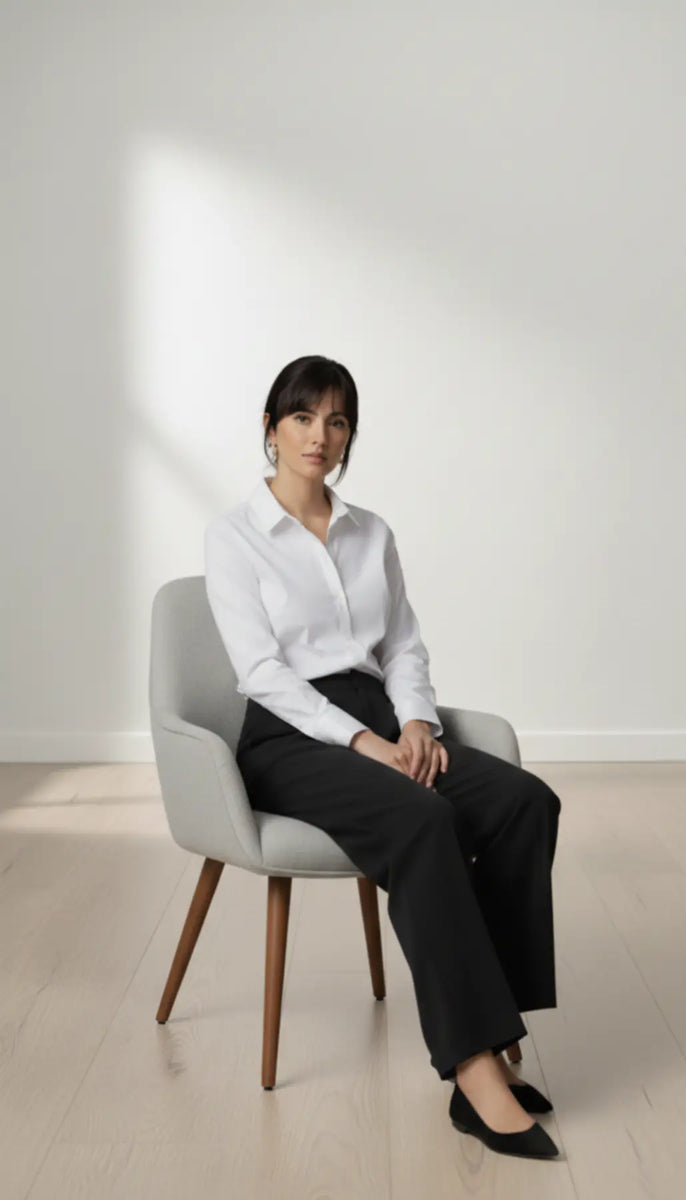 Person sitting on a chair wearing a white shirt and black trouser in a minimalistic setting.