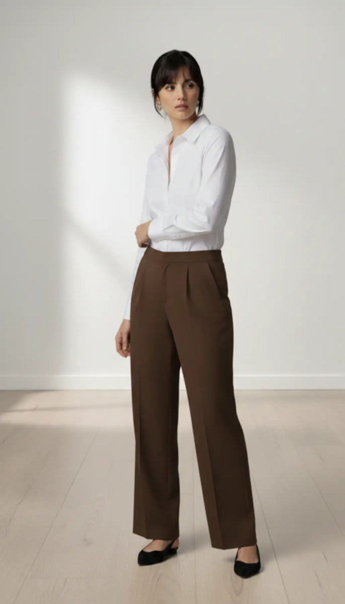 Woman wearing a white blouse and mocha trouser standing against a white wall.
