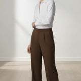 Woman wearing a white blouse and mocha trouser standing against a white wall.