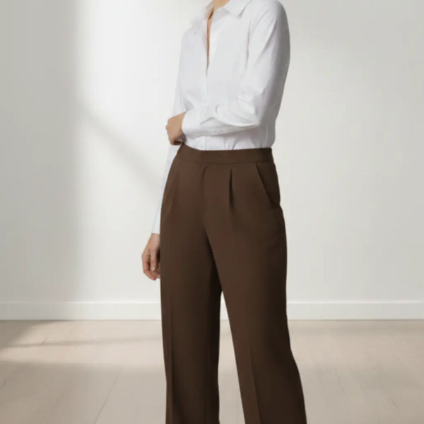 Woman wearing a white blouse and mocha trouser standing against a white wall.
