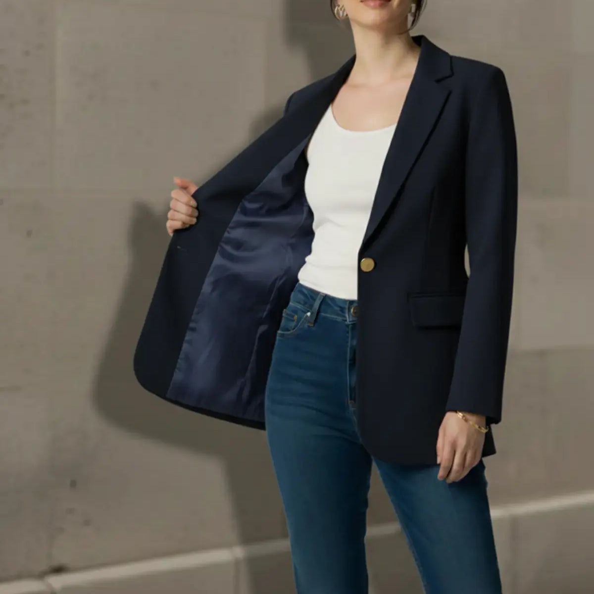 Woman wearing a navy blazer, white top, and blue jeans against a gray wall.