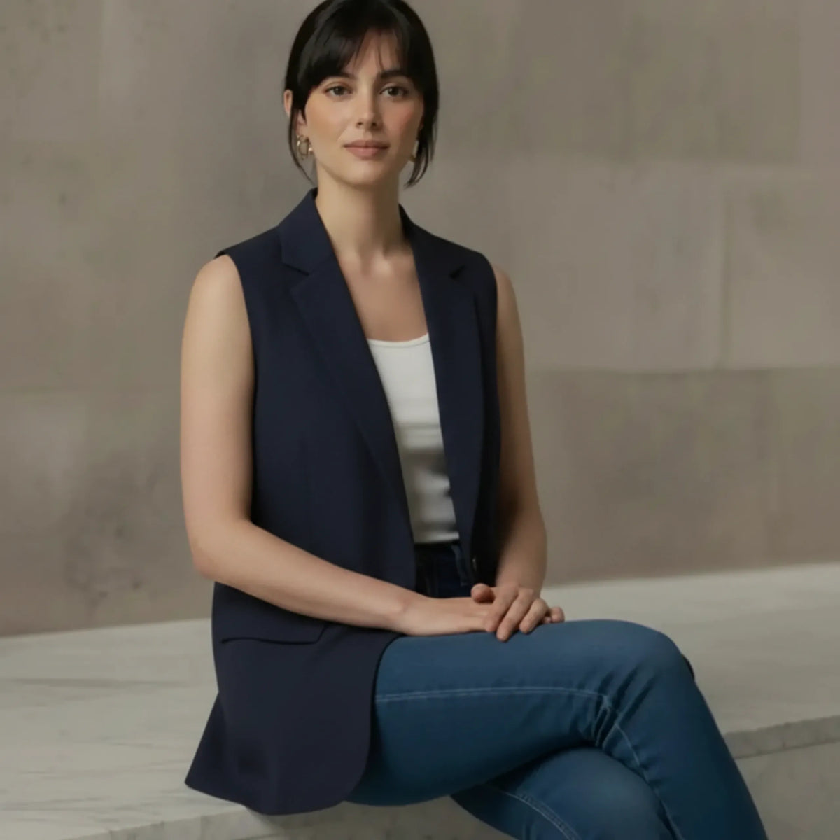 Woman sitting casually wearing an open navy blue sleeveless long vest paired with a white top and denim jeans