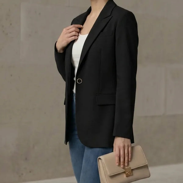 Woman in a black blazer and blue jeans holding a beige clutch against a neutral background