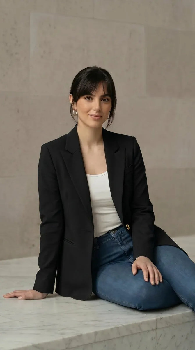 Woman wearing a black blazer and blue jeans sitting on a marble floor.