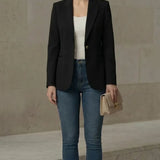 Woman wearing a black blazer, white top, blue jeans, and beige shoes against a neutral background.