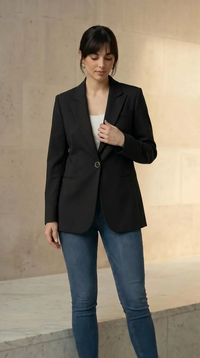 Woman wearing a black blazer and blue jeans with a beige handbag against a neutral background