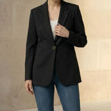 Woman wearing a black blazer and blue jeans with a beige handbag against a neutral background