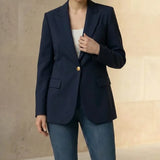 Woman wearing a navy blazer and blue jeans with a beige handbag against a neutral background