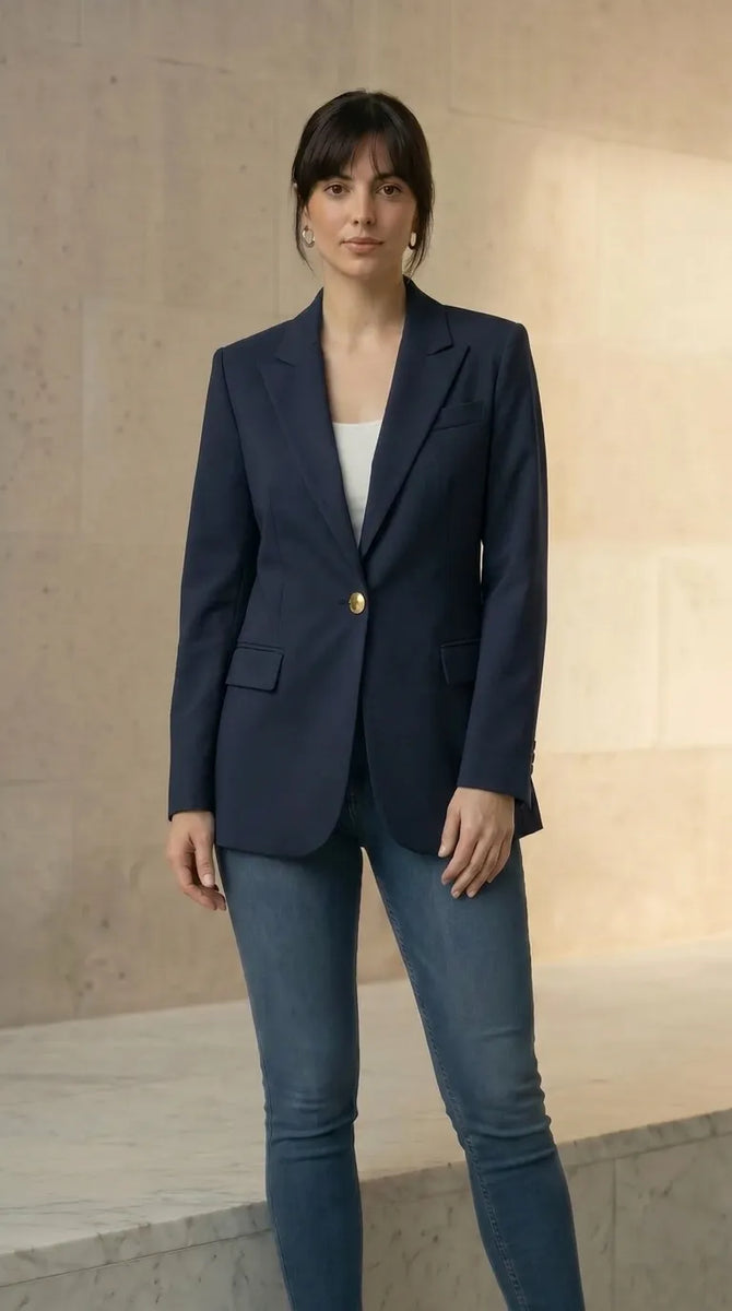 Woman wearing a navy blazer with gold buttons and blue jeans against a neutral background