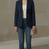 Woman wearing a navy blazer, white top, blue jeans, and beige heels against a neutral wall.