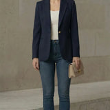 Woman wearing a navy blazer, white top, blue jeans, and beige heels against a neutral background.