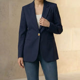 Woman wearing a navy blazer with gold buttons and blue jeans, standing against a neutral background.