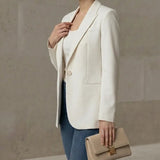 Woman wearing a white blazer and blue jeans, holding a beige clutch against a neutral background