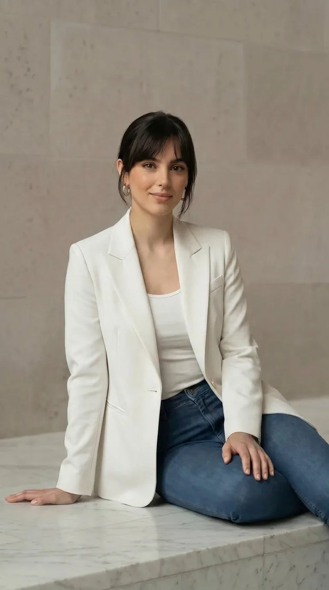 Woman wearing a white blazer and blue jeans sitting on a marble ledge.