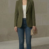 Woman wearing an olive green blazer, white top, blue jeans, and beige heels against a neutral background.