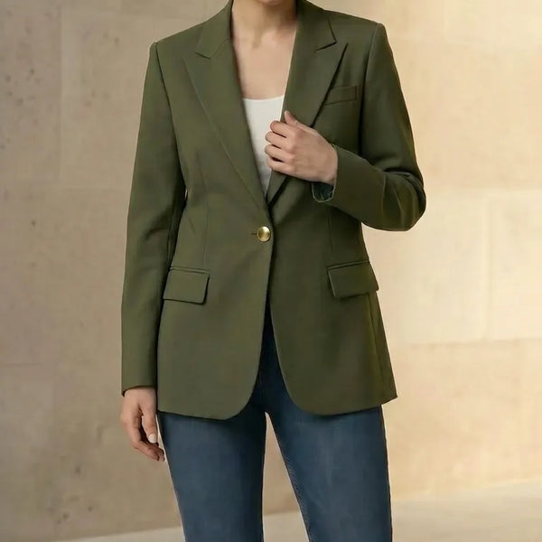 Woman wearing an olive blazer and blue jeans, holding a beige handbag against a neutral background.