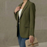 Woman wearing an olive green blazer and blue jeans, holding a beige clutch against a neutral background.